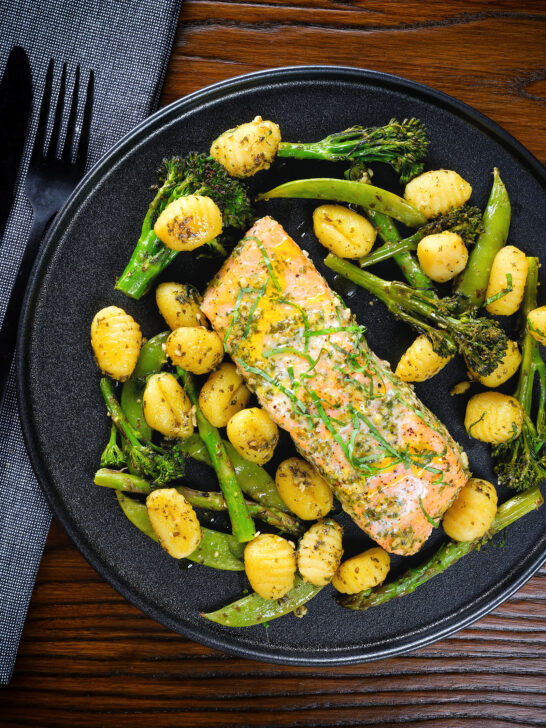 Plateau de filet de saumon cuit au four avec pesto, légumes verts et gnocchis servis sur une assiette noire.