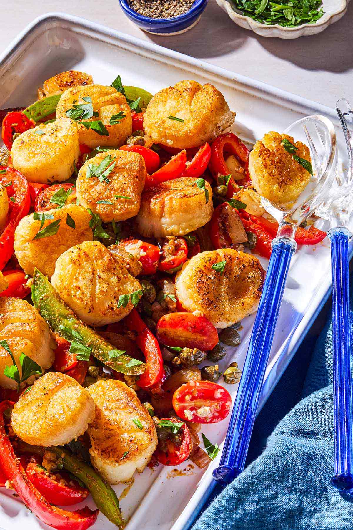 Pétoncles méditerranéens nichés avec des tomates et des poivrons sur un plateau de service avec ustensiles de service.