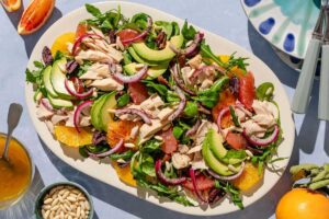 Avocado tuna salad on a platter surrounded by a stack of 2 plates with forks, orange wedges, salad dressing, and a bowl of pine nuts.