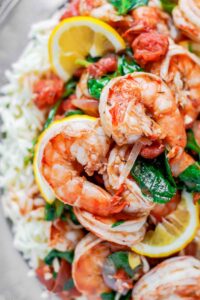 close up of two shrimp with orzo, wilted spinach, and lemon wedges in the background.