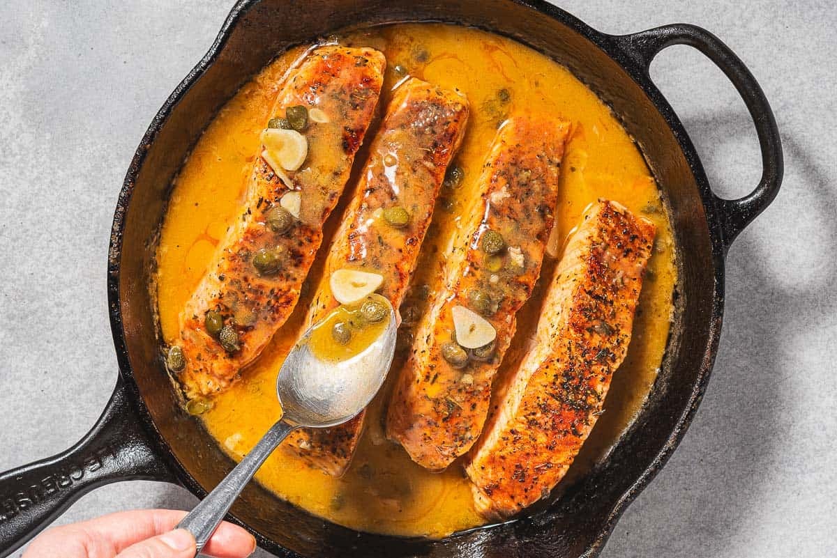 Sauce en cuillère sur les 4 filets de piccata de saumon dans une poêle en fonte.