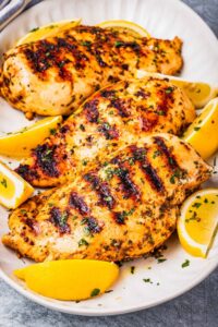 Three grilled chicken breasts that have been marinated in Greek chicken marinade and are on a platter with lemon wedges and a sprinkle of parsley.