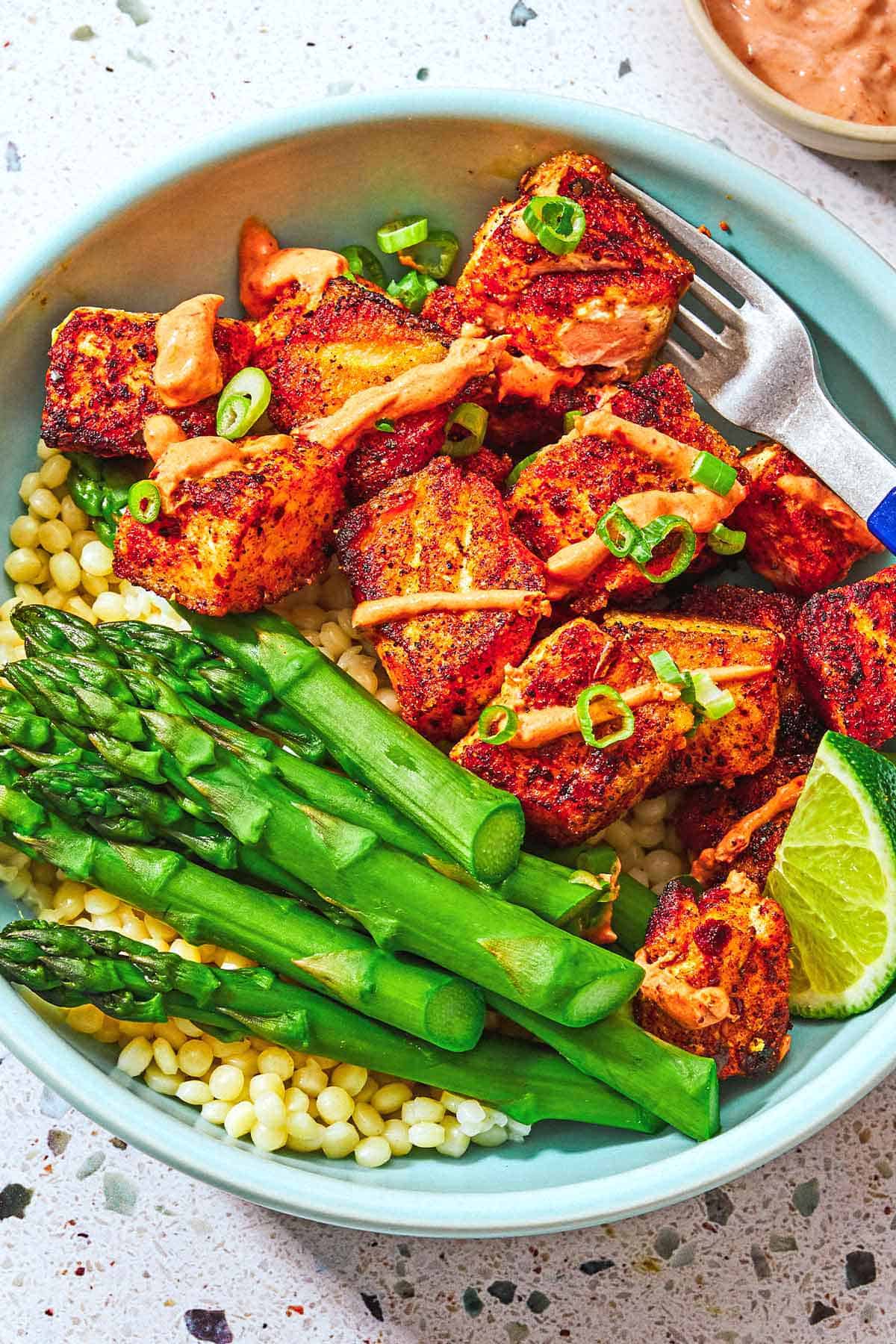 Plusieurs bouchées de saumon croustillantes garnies d'un filet de sauce au yaourt de Harissa et d'oignons verts et d'asperges sur un lit de couscous perlé dans un bol avec une fourchette.