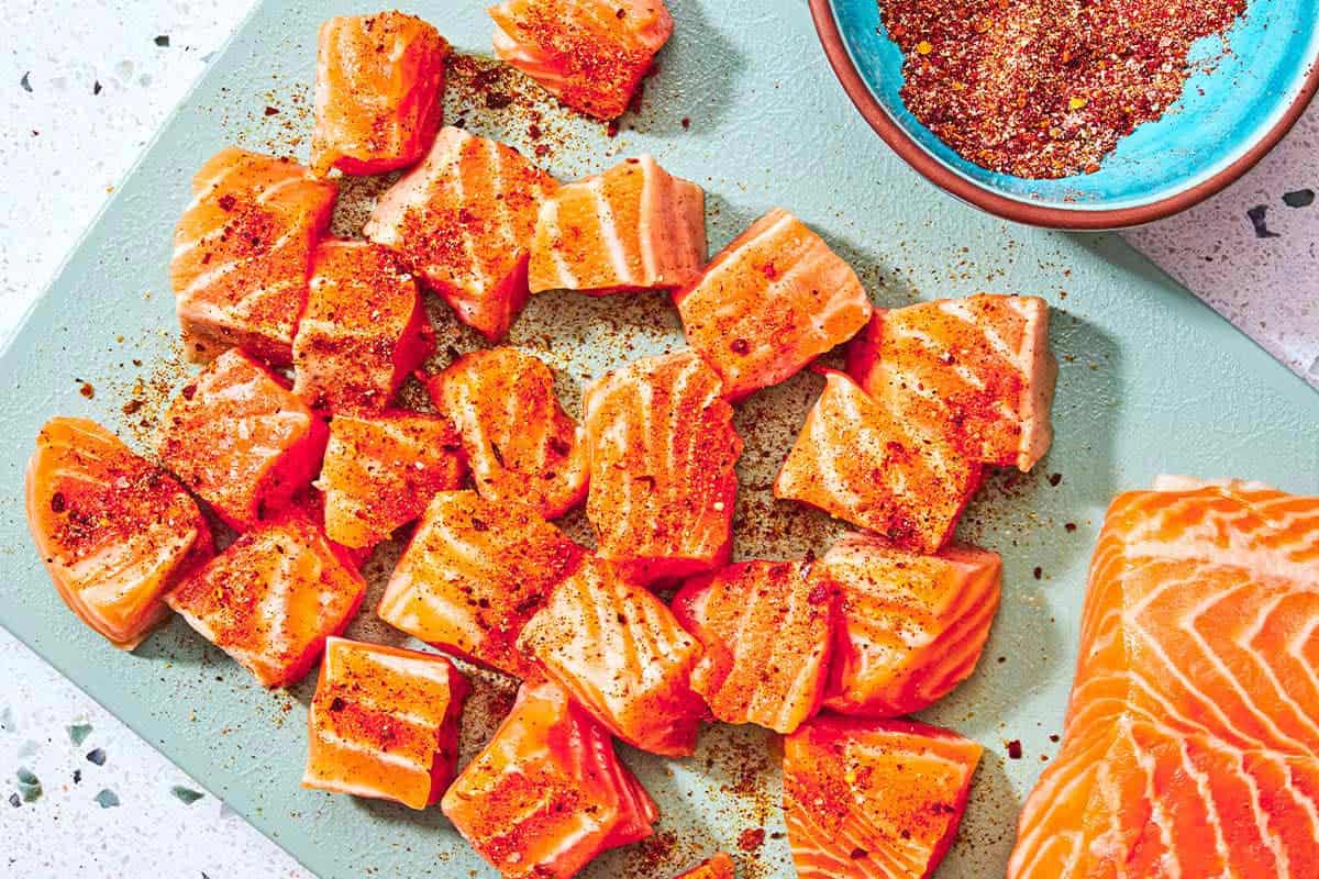 Les piqûres de saumon assaisonnées non cuites sur une planche à découper avec le reste du filet de saumon à côté d'un bol d'assaisonnement.