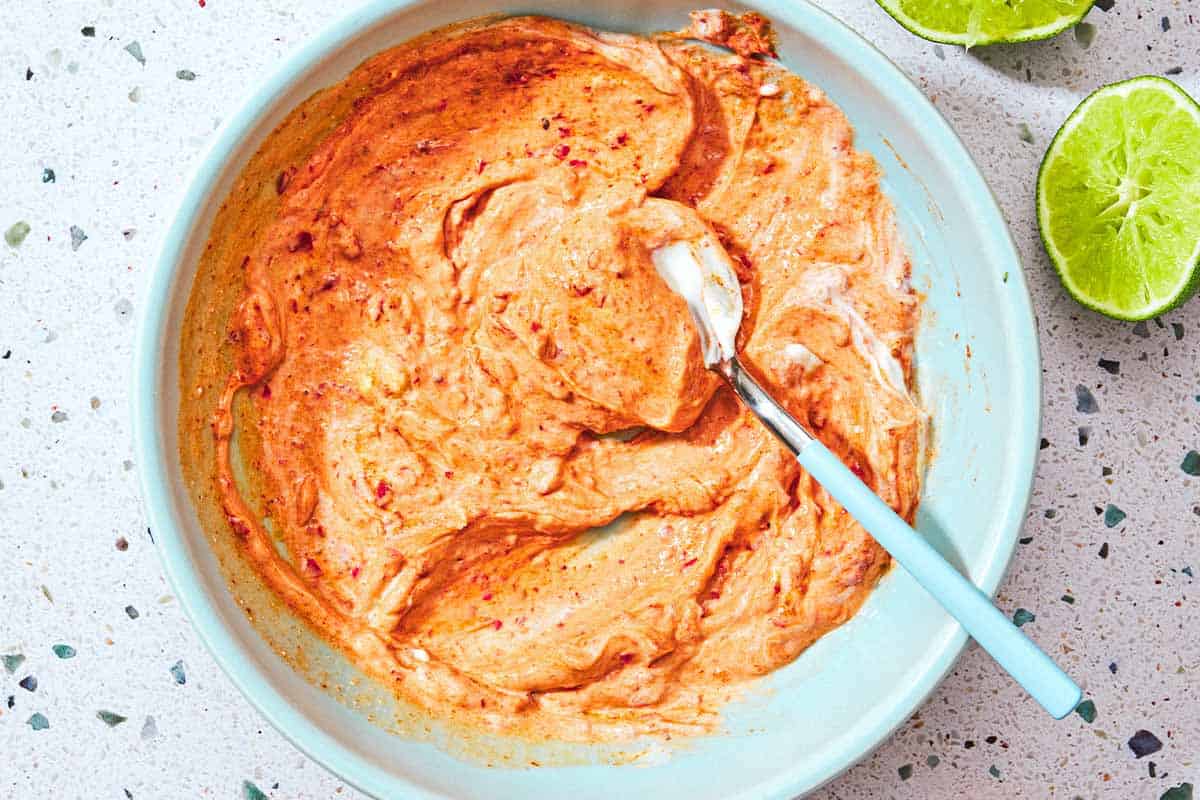 La sauce harissa-yogourt pour les piqûres de saumon dans un bol avec une cuillère à côté de deux moitiés au citron vert.