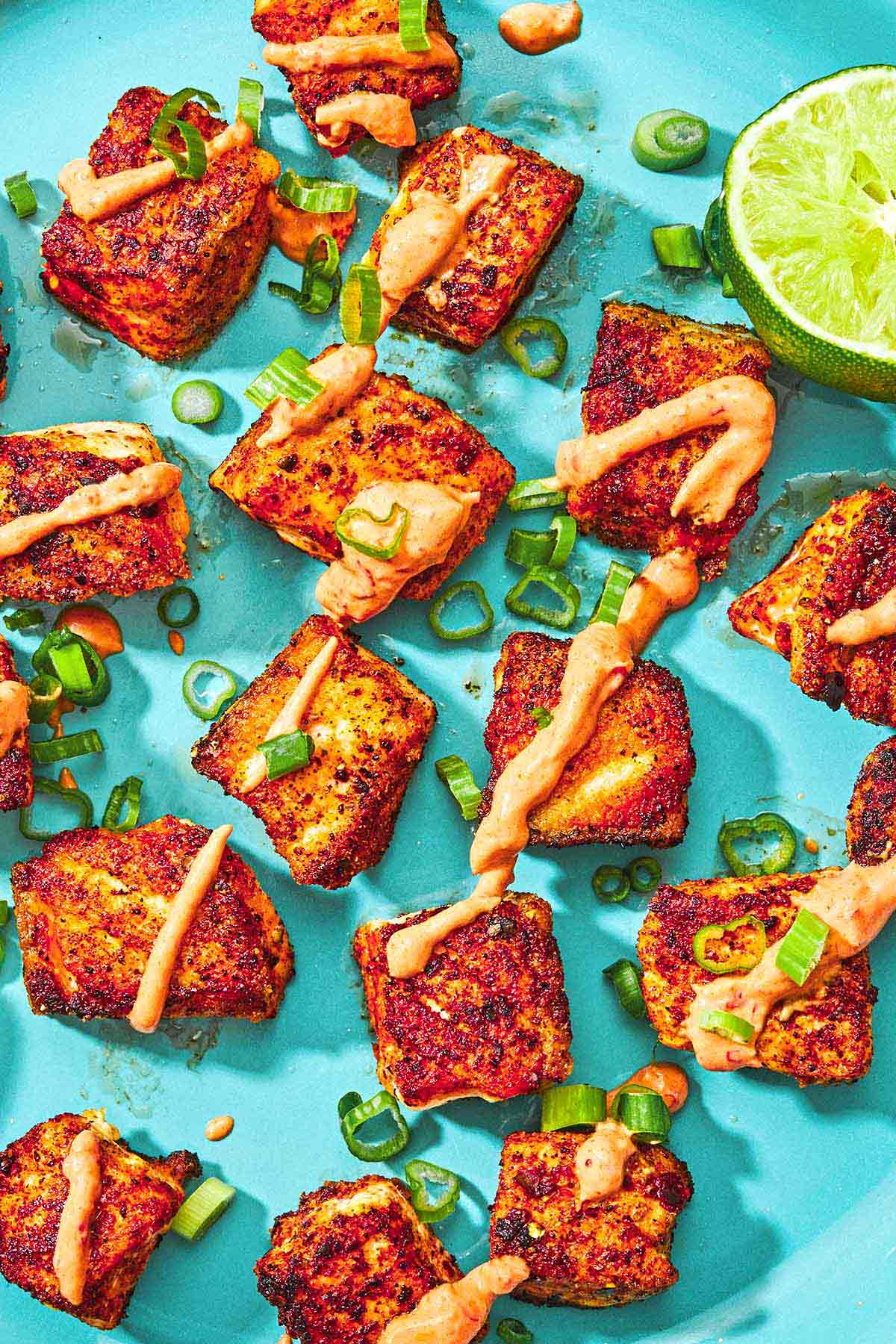 Plusieurs bouchées de saumon croustillantes garnies d'un filet de sauce au yaourt de Harissa et d'oignons verts sur une assiette avec une moitié de citron à jus.