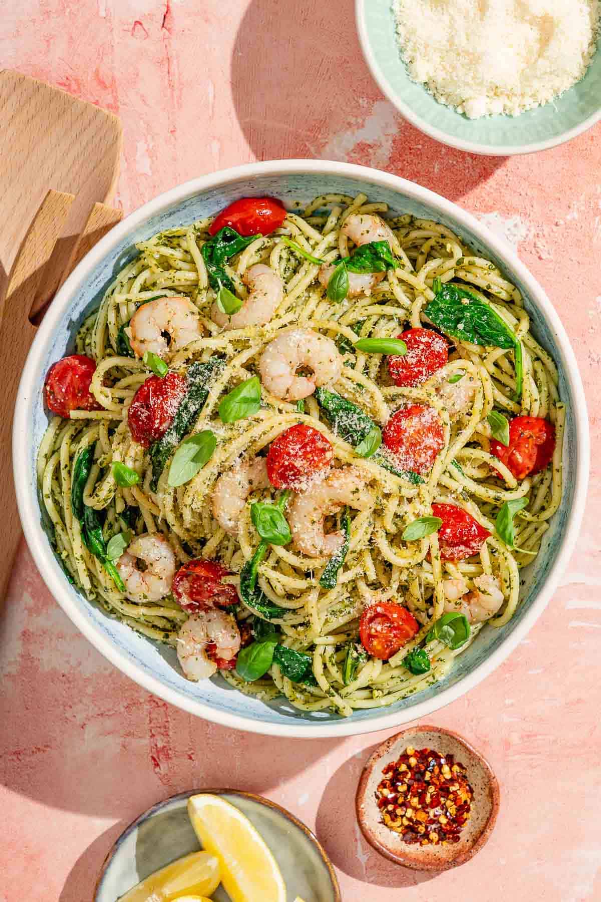 Pâtes de pesto de crevettes dans un bol de service entouré d'un bol de parmesan râpé, d'une assiette de quartiers de citron et d'un petit bol de flocons de poivron rouge.