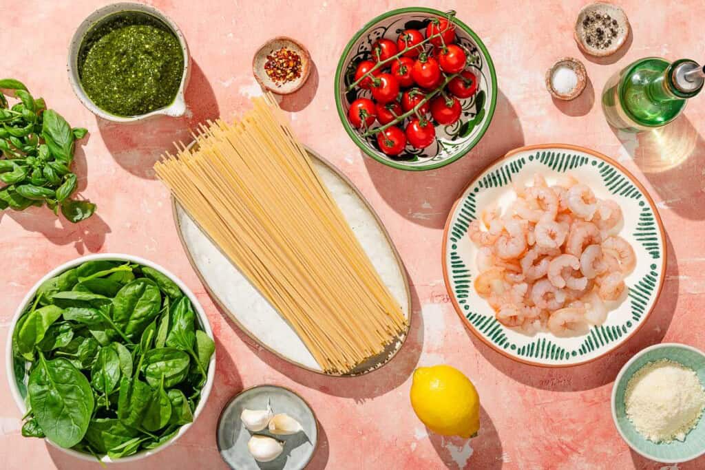 Ingrédients pour les pâtes au pesto de crevettes, y compris les crevettes, les spaghettis, le sel, le poivre noir, le bicarbonate de soude, l'huile d'olive, l'ail, les tomates cerises, le pesto, les flocons de poivron rouge, les bébés épinards, le parmesan râpé et le basilic.