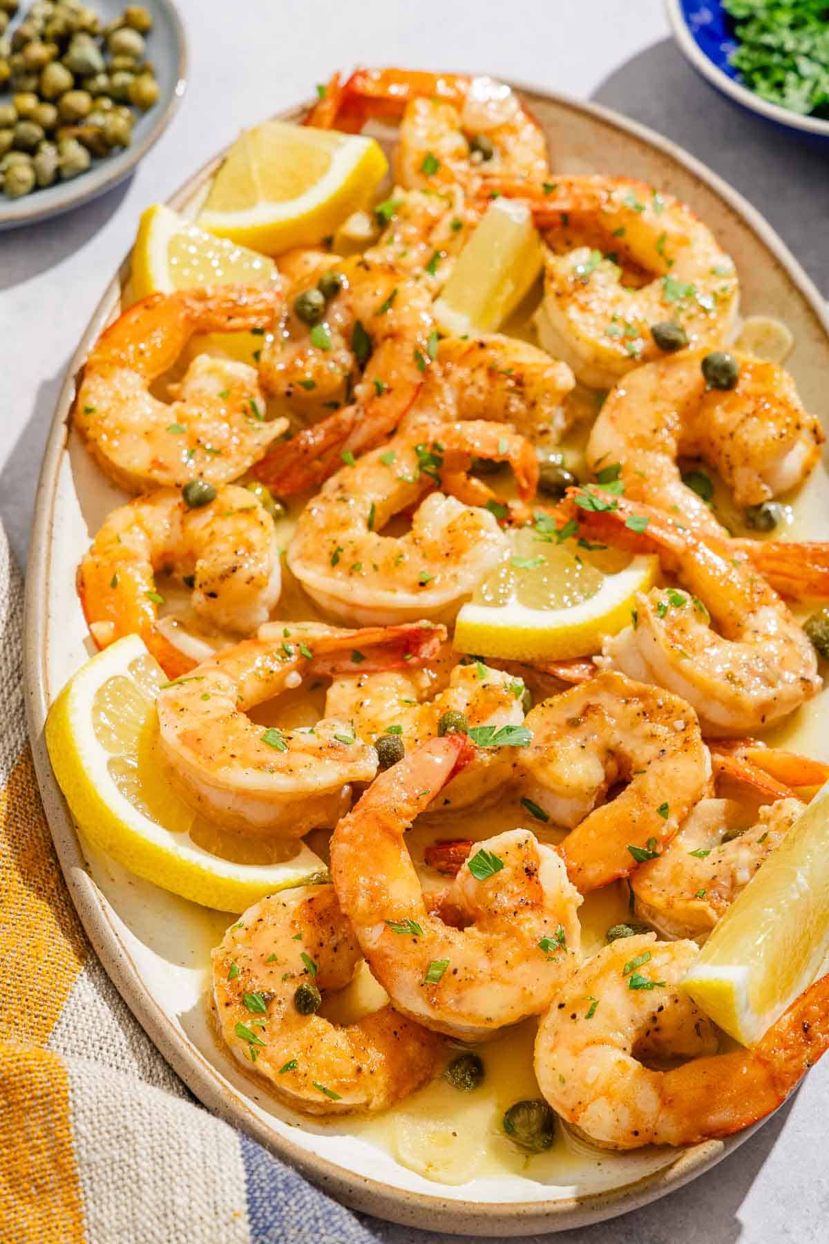 Piccata de crevettes avec des quartiers de citron sur un plateau à côté de petits bols de persil haché et de câpres.