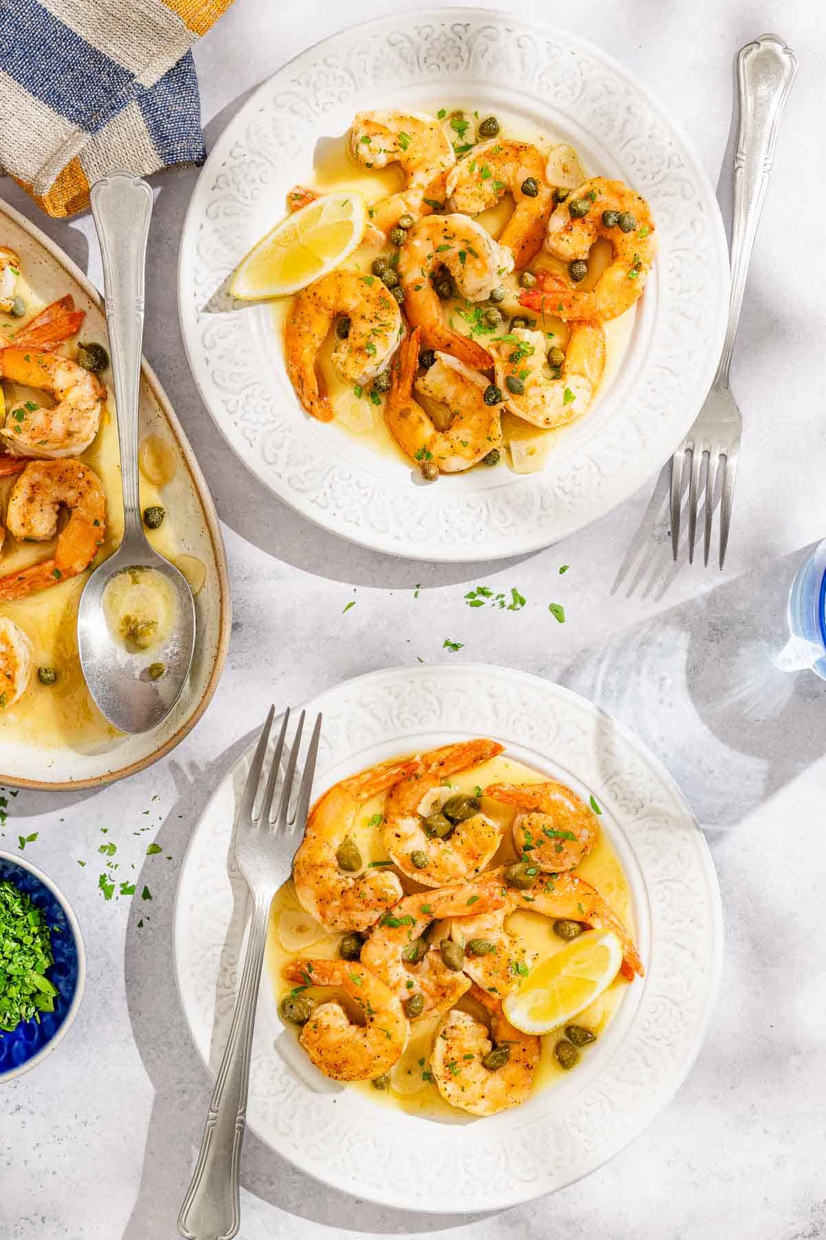 Deux portions de piccata de crevettes avec des quartiers de citron sur des assiettes avec des fourches. À côté de cela se trouve le reste du Piccata de crevettes sur un plateau avec une cuillère en service et un bol de persil haché.