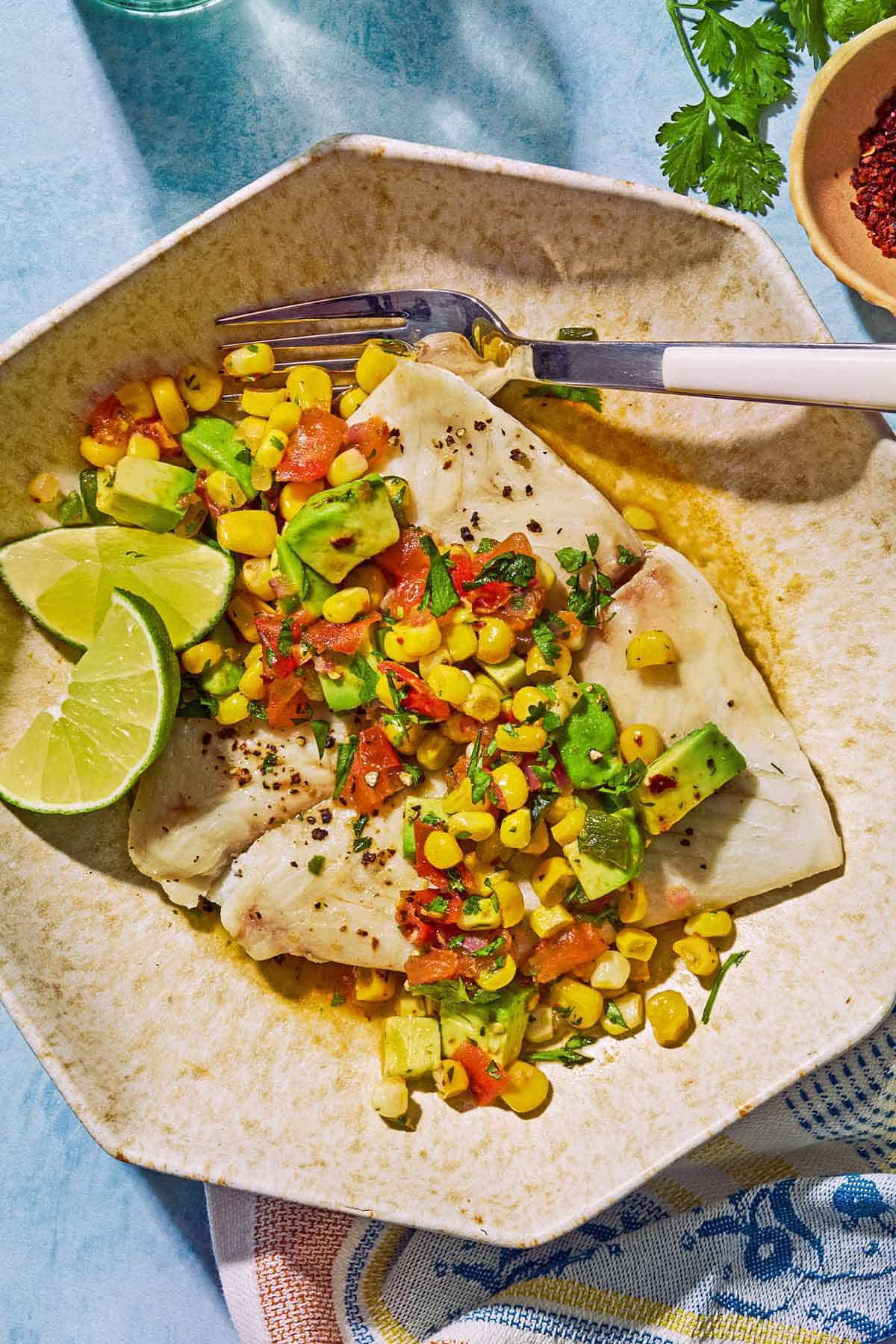 Pole pochée garnie de salsa de maïs sucré sur une assiette avec des quartiers de citron vert et une fourchette. À côté de cela se trouve un torchon, un bol de poivre d'Alep et quelques brins de coriandre.