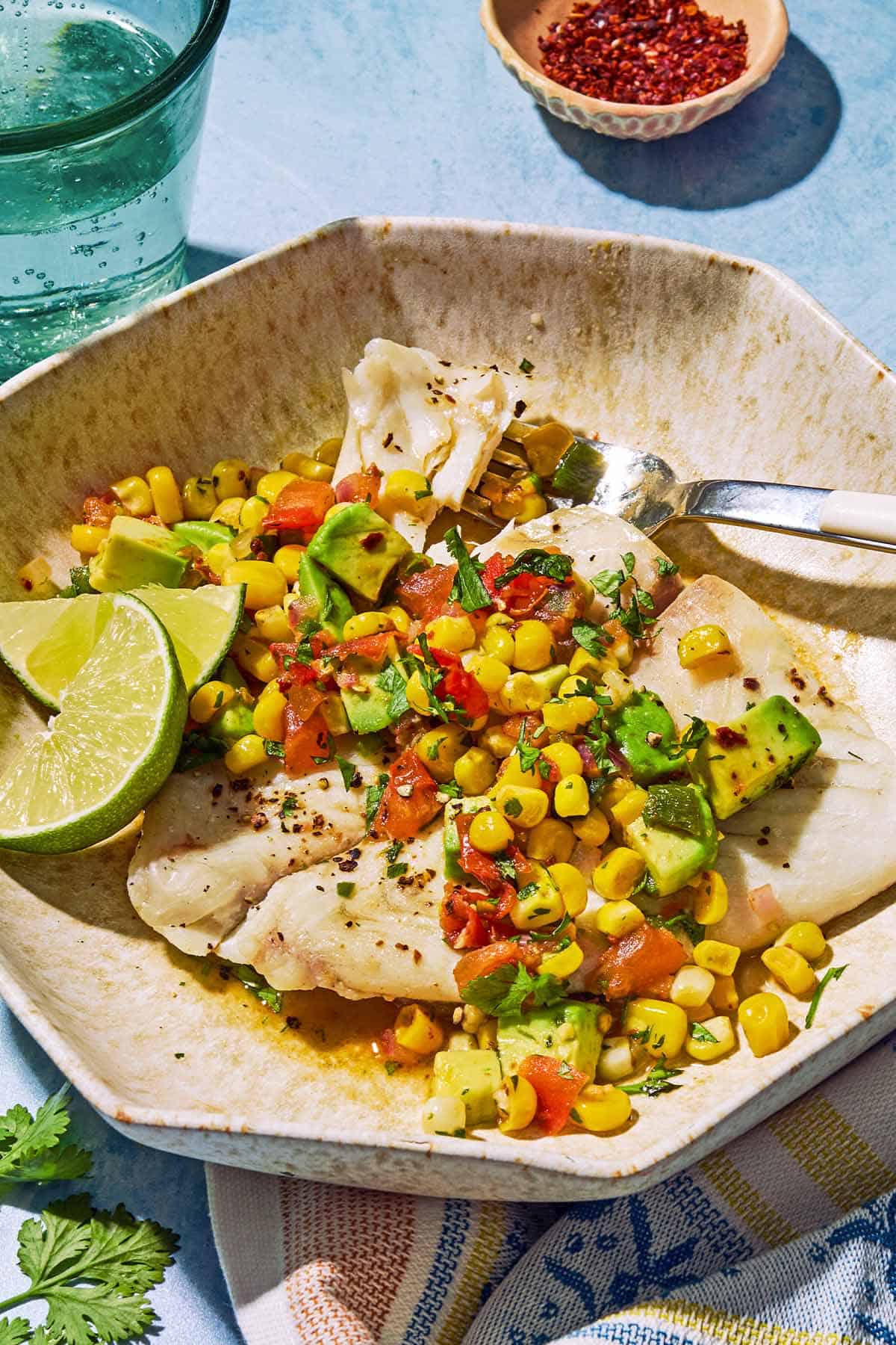 Pole pochée garnie de salsa de maïs sucré sur une assiette avec des quartiers de citron vert et une fourchette. À côté de cela se trouve un torchon, un bol de poivre d'Alep et un verre d'eau mousseuse.