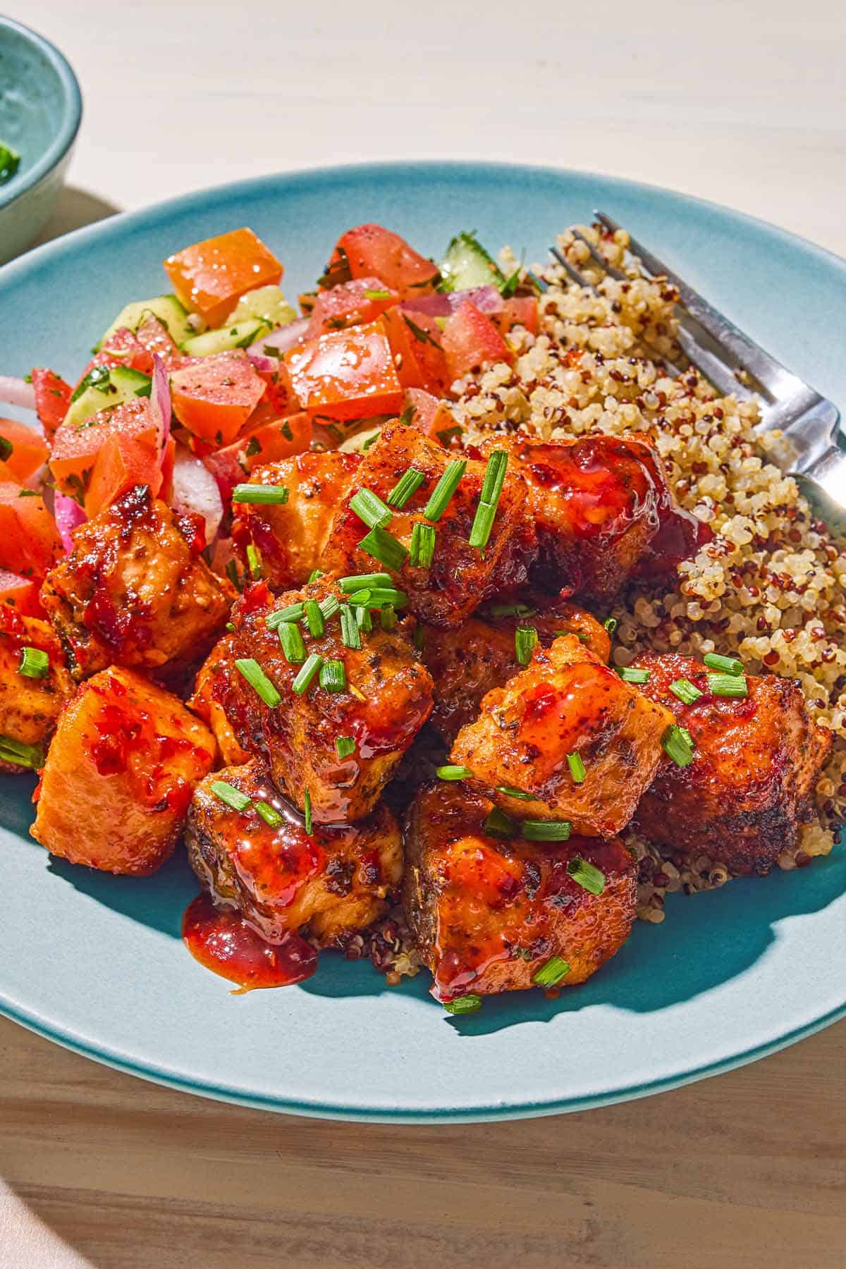 Une portion de piqûres de saumon de friteuse à air garni de ciboulette hachée dans une assiette avec du quinoa, de la salade de concombre de tomate et une fourchette.