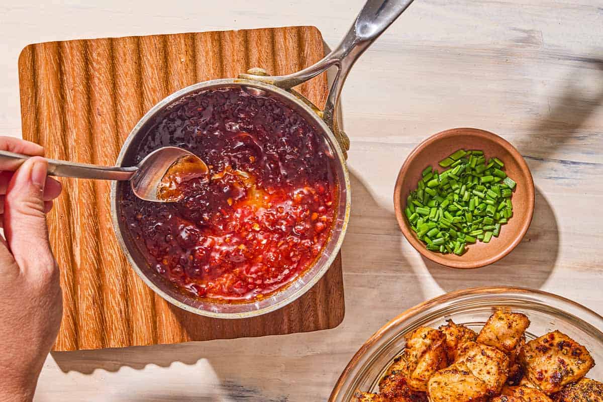 Le glaçage au miel-harissa étant agité dans une casserole sur un trivassement en bois. À côté de cela se trouve un petit bol de ciboulette hachée et un bol de piqûres de saumon à air.