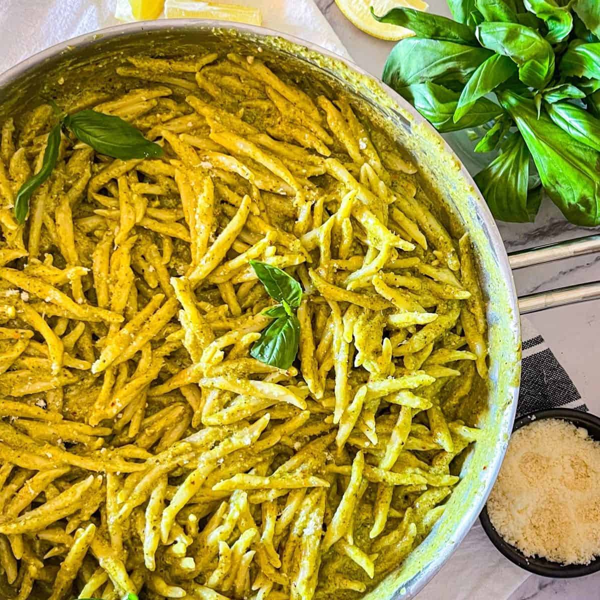 Pesto pasta in a skillet with basil and cheese next to it.