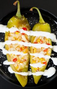 A small black plate with two large hatch peppers that have been stuffed with crab, corn, red bell pepper, and cheese. They ache been drizzled with lime crema and sprinkled with cojita cheese.