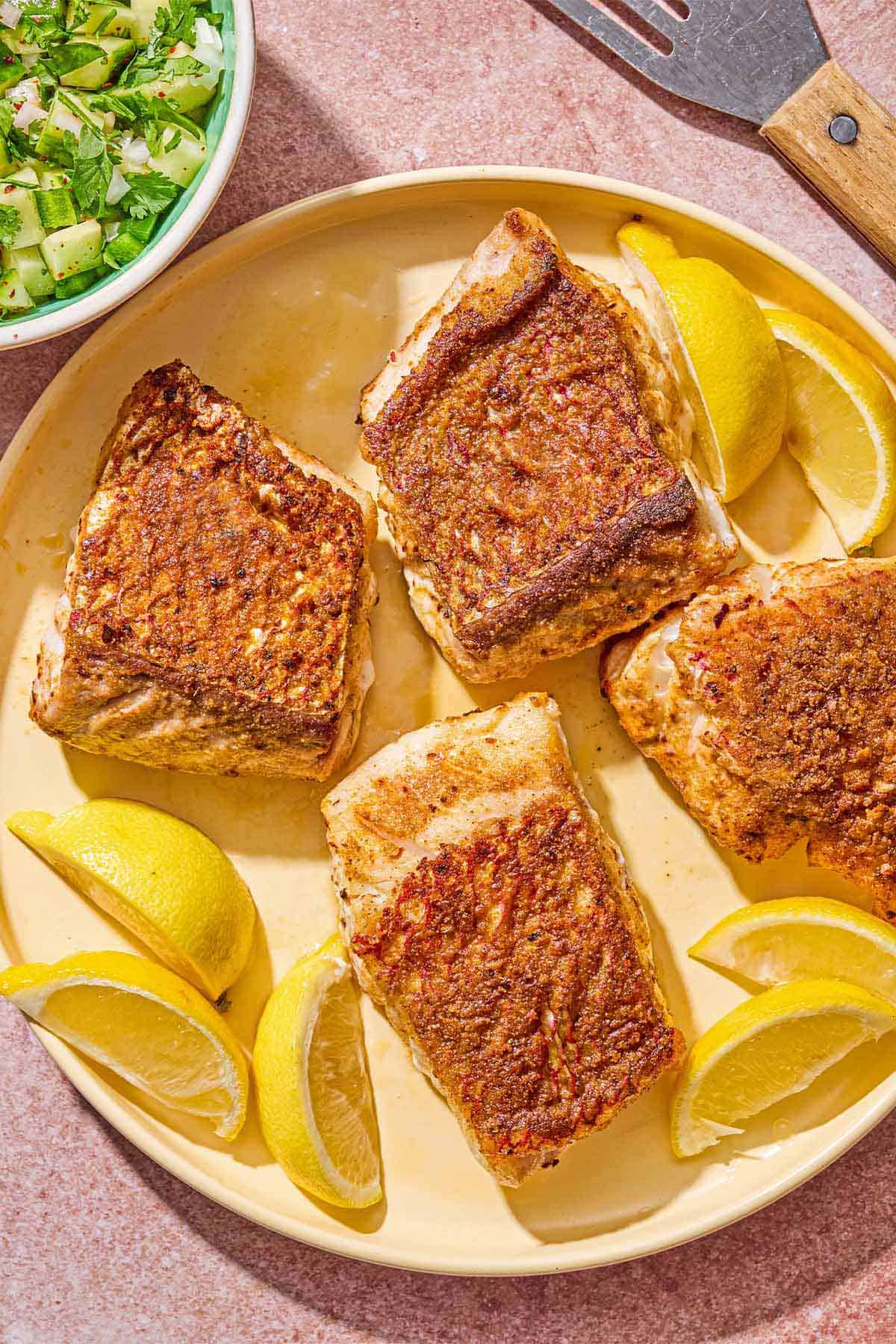 4 filets de vivaneau frits sur une assiette avec des quartiers de citron. À côté se trouvent un bol de salsa de concombre et une spatule.