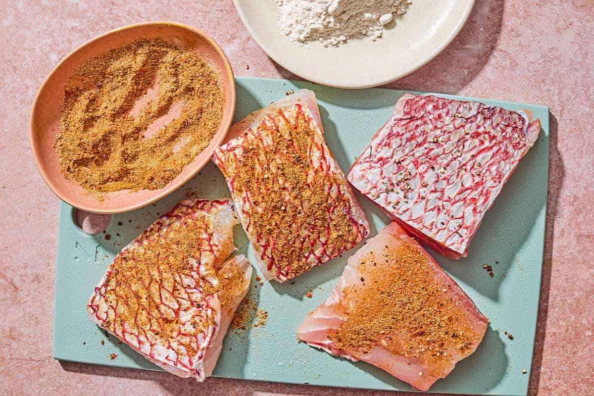 Quatre filets de vivaneau sur une planche à découper avec un bol d'assaisonnement. L'assaisonnement a été ajouté à 3 des filets.