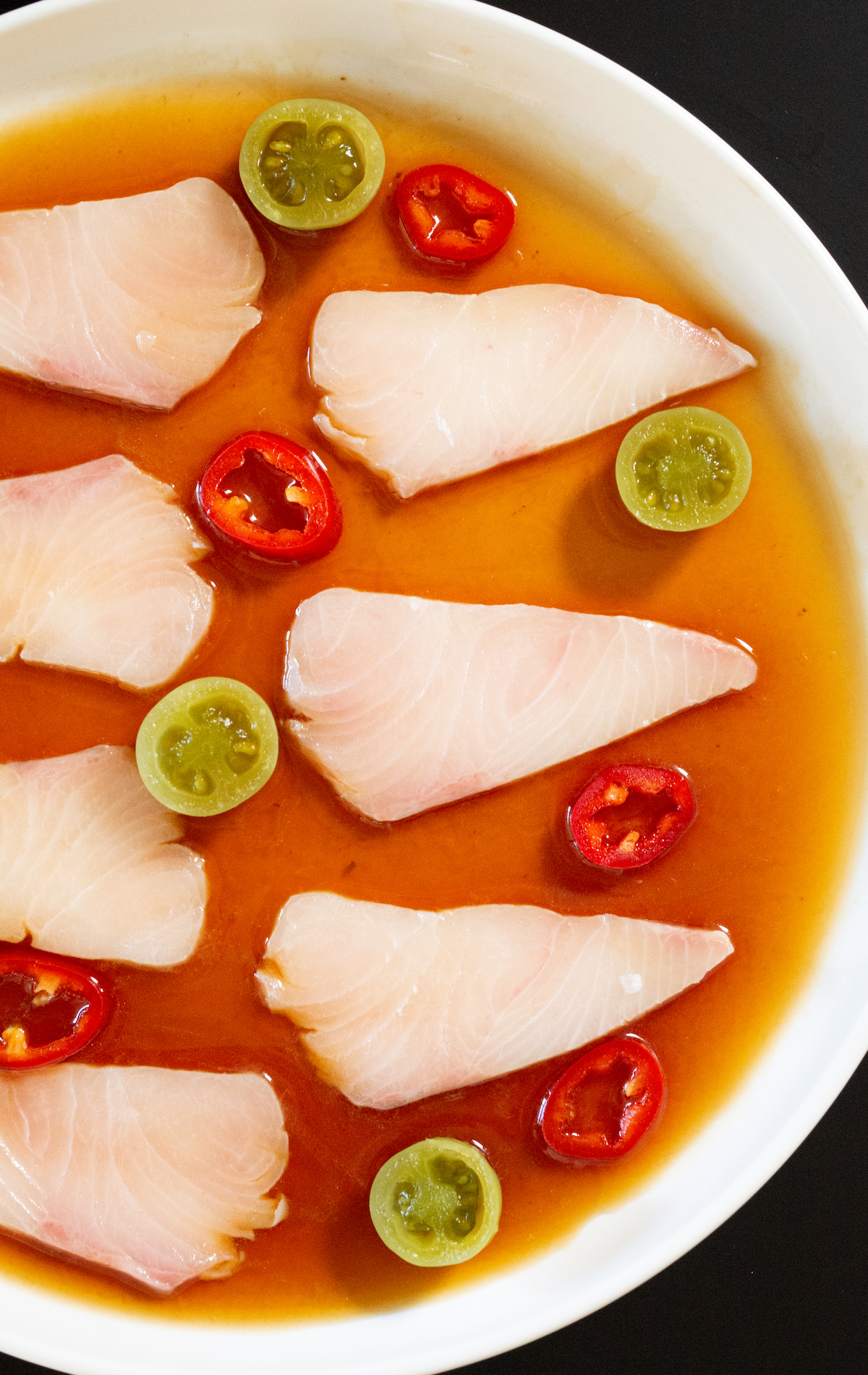Une assiette blanche remplie d'une sauce brun rougeâtre. Des tranches de hamachi cru sont posées sur le dessus, à côté de tomates vertes marinées coupées en deux et de tranches de piment Fresno rouge.