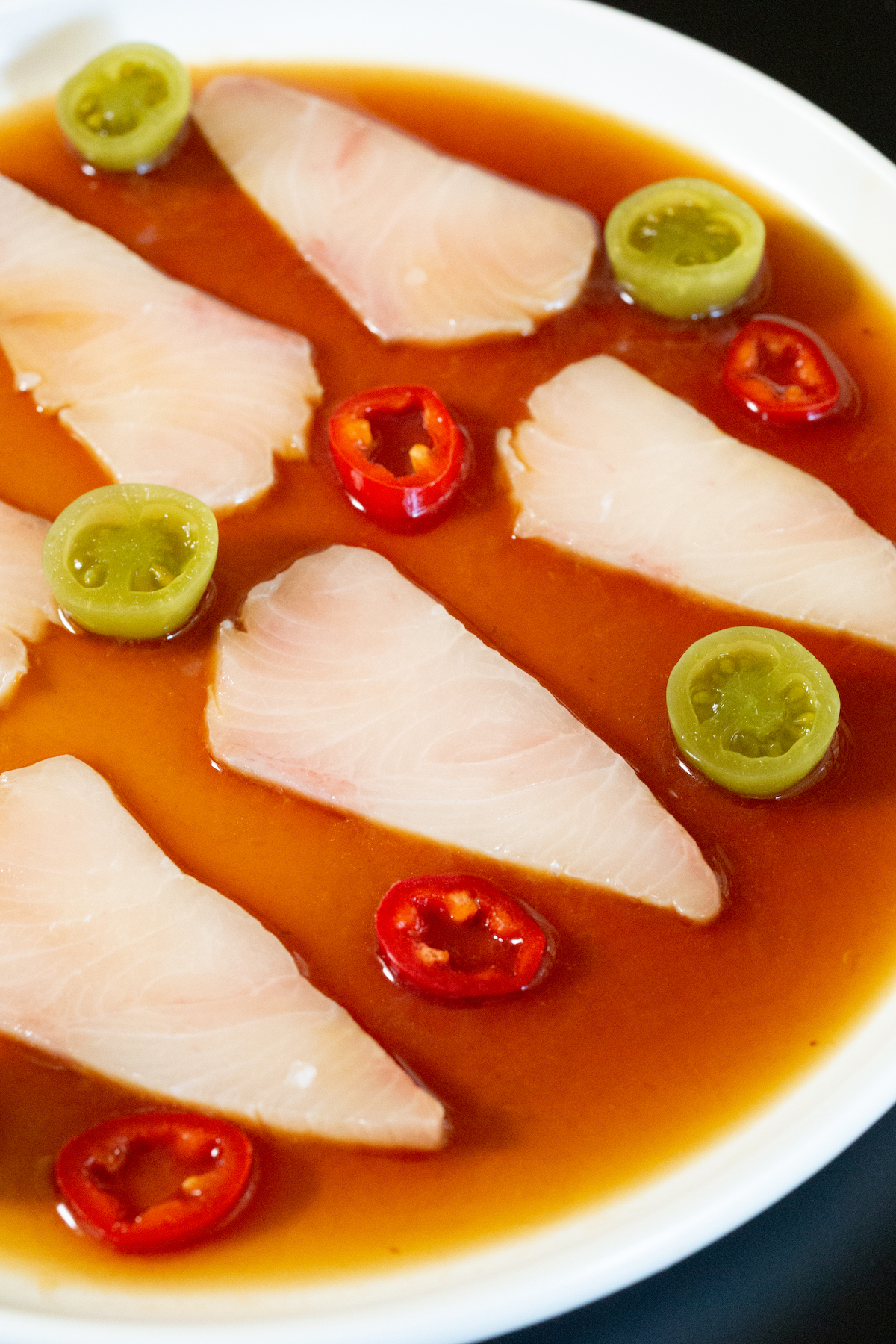 Plusieurs tranches de hamachi cru sur une assiette remplie d'une sauce brun rougeâtre. Des tranches de piments Fresno et des tomates vertes marinées sont disposées autour des tranches de poisson.