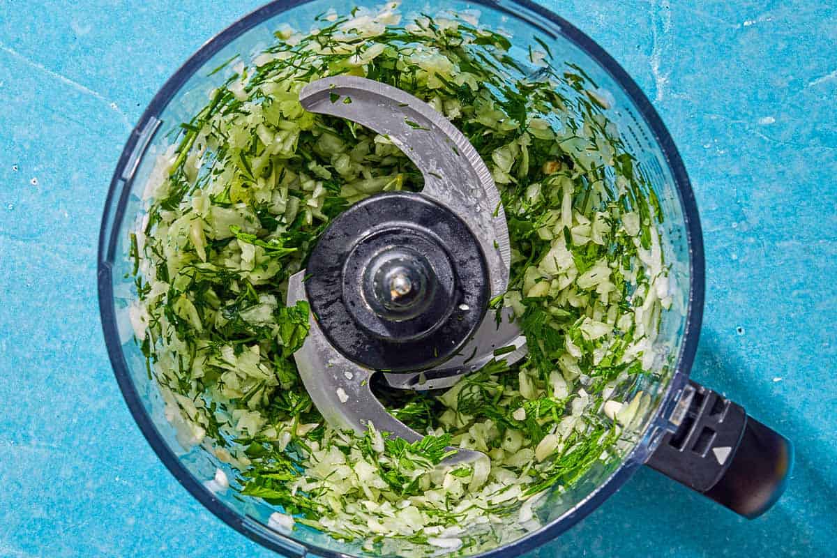 L'oignon, le persil, l'aneth et l'ail dans le bol d'un robot culinaire juste après avoir été mélangés.