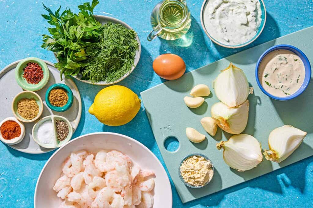 Ingrédients pour la kofta aux crevettes comprenant les crevettes, l'oignon, le persil, la garniture, l'ail, le sel, le poivre, la coriandre, le piment d'Alep, le cumin, le paprika, la farine de pois chiches, l'œuf, l'huile, le citron, la sauce tahini et la sauce tzatziki.