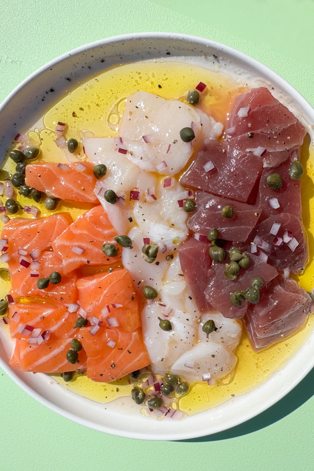 A small white serving plate is filled with olive oil and lemon juice with raw scallop slices, salmon pieces, and tuna pieces sitting on top. It