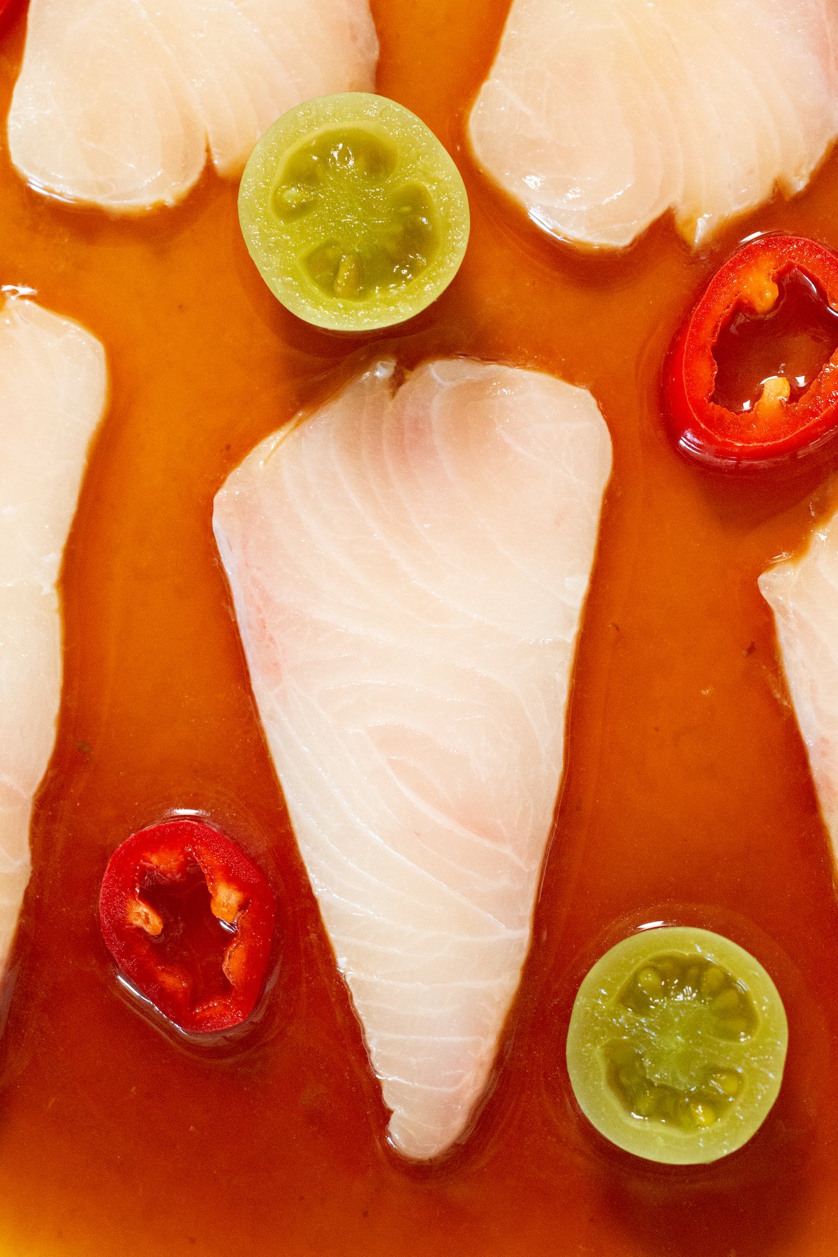 Gros plan d'une tranche de hamachi où l'on peut clairement voir les marbrures du poisson. Il est servi dans une sauce brun-rouge à côté de tomates vertes coupées en deux et de tranches de piment rouge.