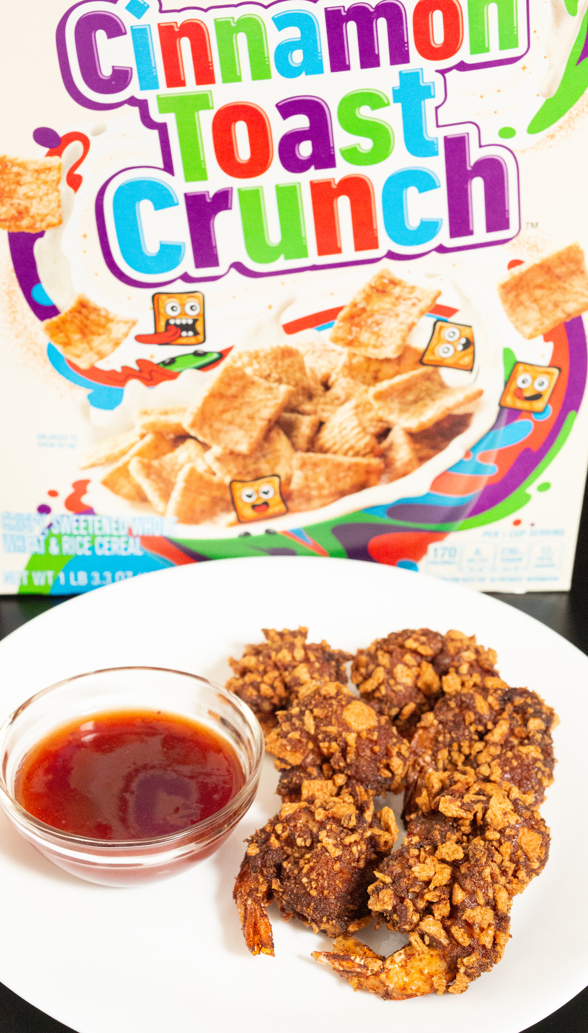 Six crevettes panées et frites Cinnamon Toast Crunch sont posées sur une assiette blanche à côté de la sauce habanero aux fraises et devant une boîte de céréales Cinnamon Toast Crunch.