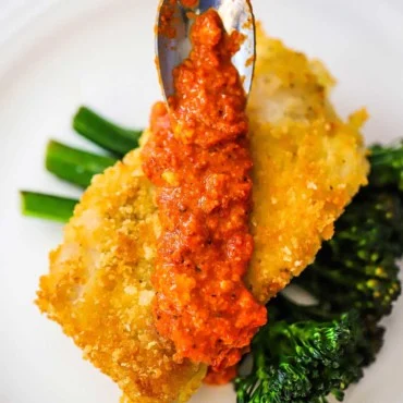 Une petite cuillère utilisée pour ajouter de la sauce romesco sur la morue en croûte de parmesan sur un bouquet de brocolini sautés.