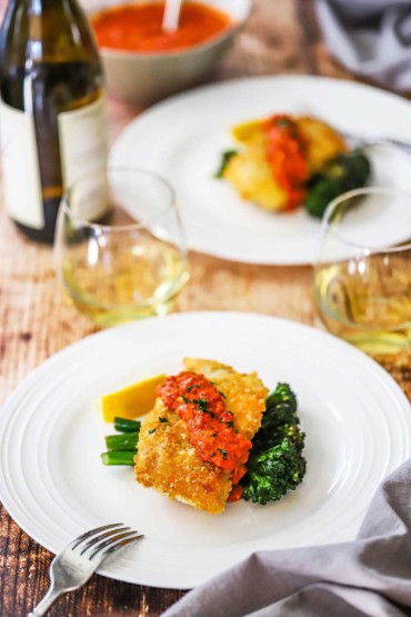 Deux assiettes remplies d'une portion de morue en croûte de parmesan avec romesco et brocolini sautés à côté de deux verres de vin blanc.