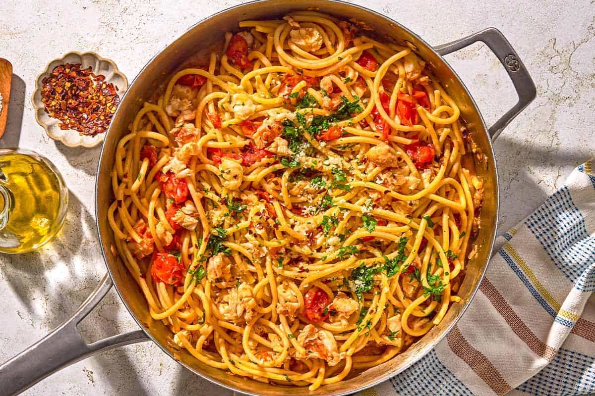 Pâtes au homard dans une grande poêle à côté d'un bol de flocons de piment rouge, d'une bouteille d'huile d'olive et d'un torchon.