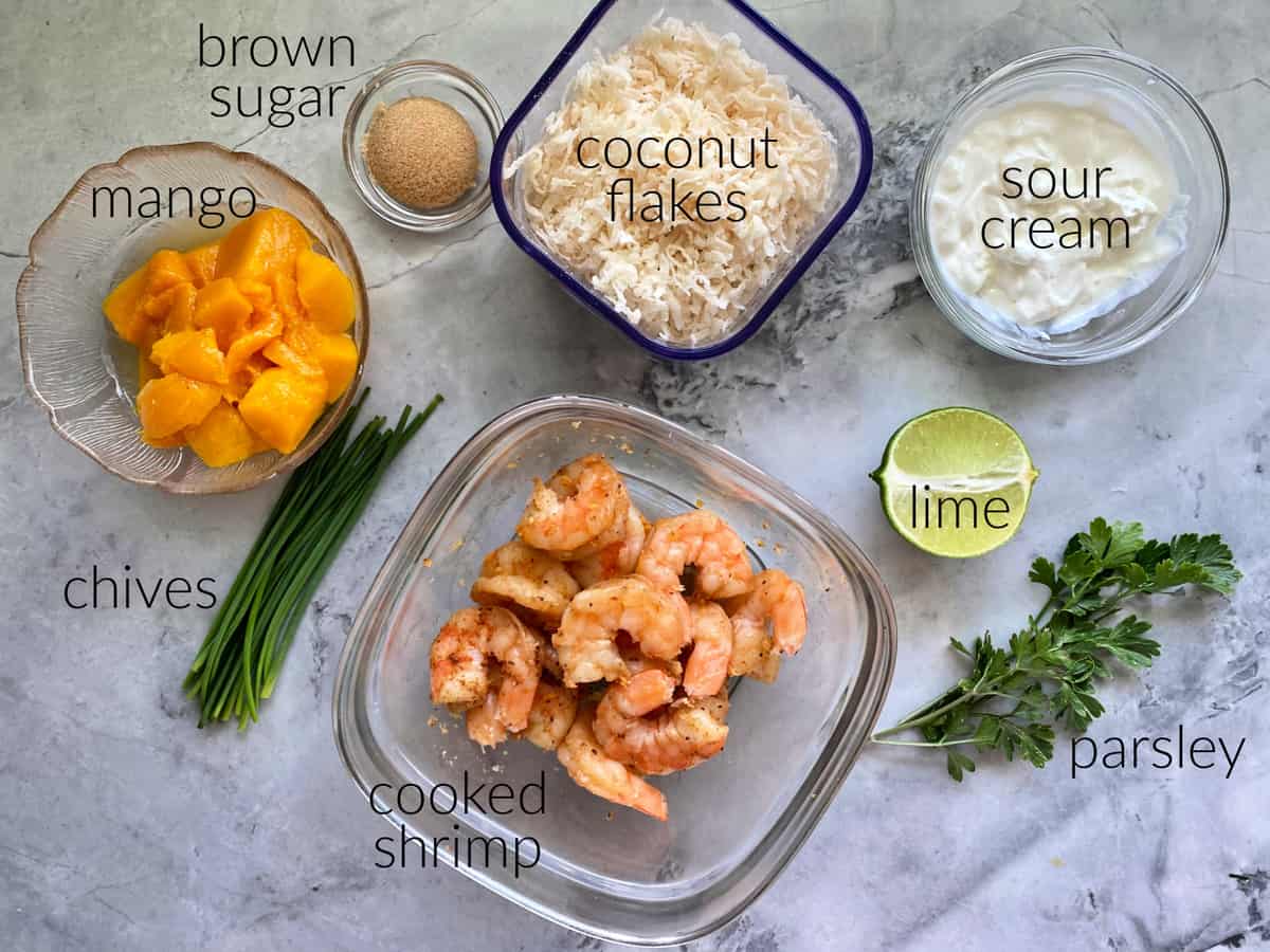 Ingrédients sur le plan de travail : mangue, cassonade, ciboulette, crevettes, flocons de noix de coco, crème sure, citron vert, persil.