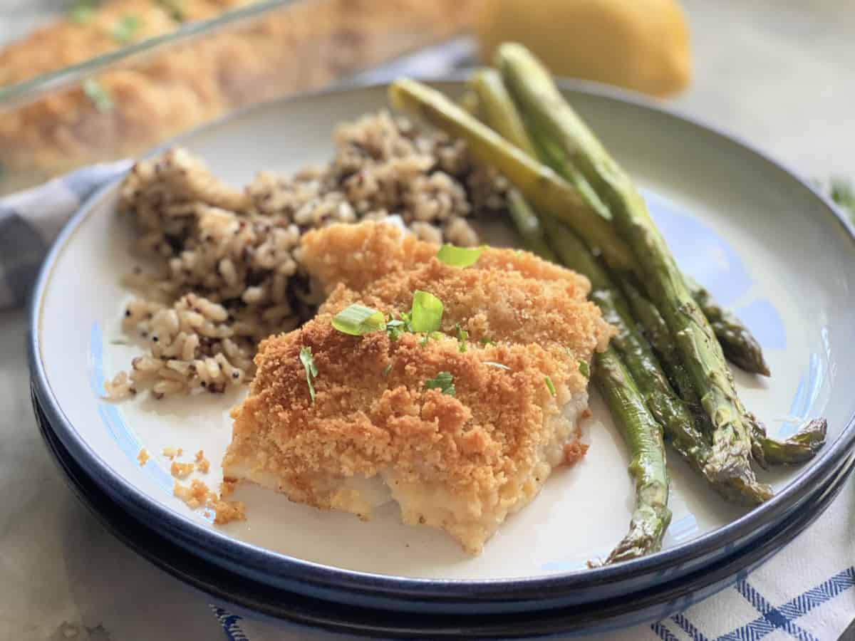 Poisson pané sur une assiette à côté d'asperges et de riz avec un plat de cuisson en verre en arrière-plan avec du poisson.