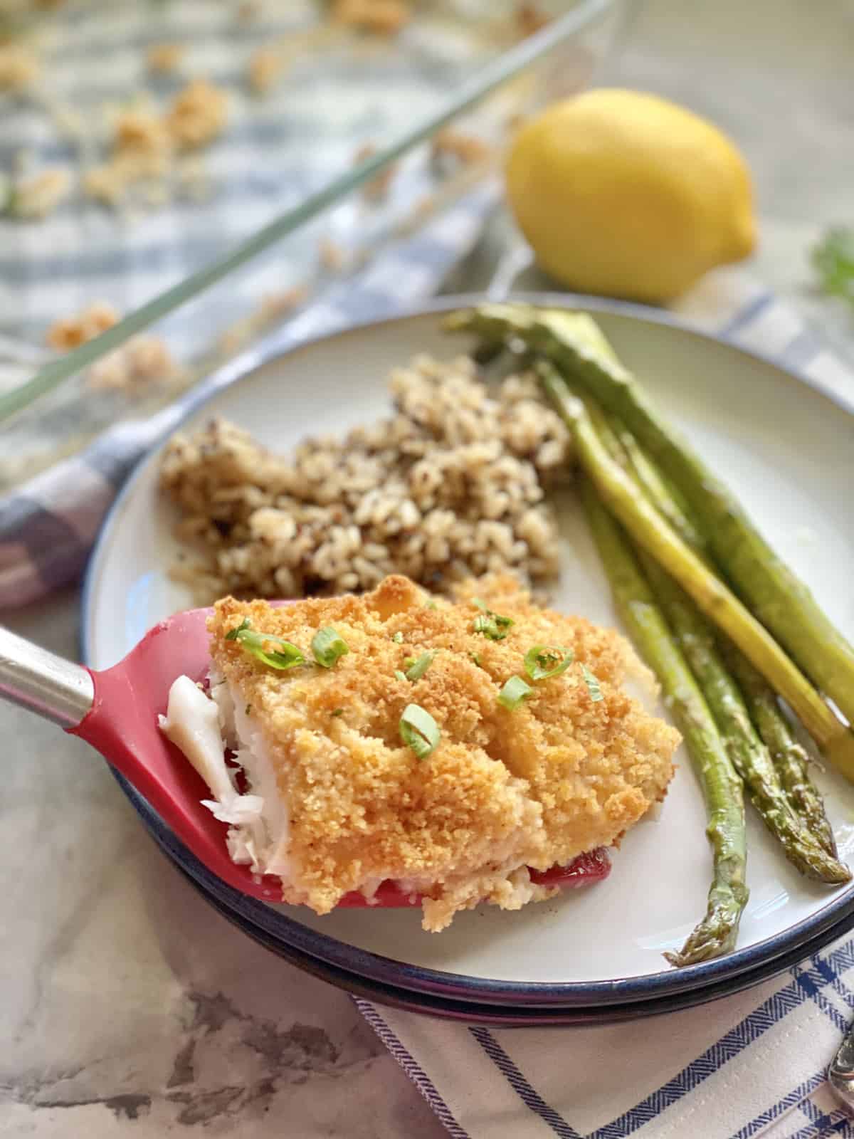 Spatule rouge plaçant un morceau de poisson pané sur une assiette avec du riz et des asperges.