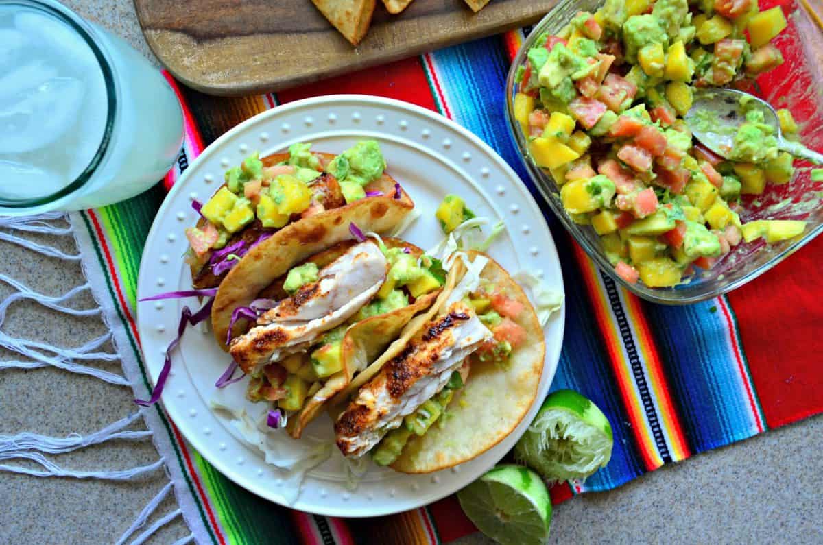 vue de dessus 4 tacos au poisson Mahi Mahi plaqués avec salsa à la mangue et à l'avocat à côté d'un bol de salsa.
