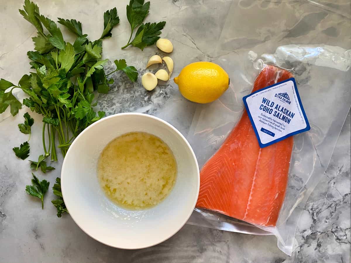 Vue de dessus du saumon emballé, du beurre fondu, des gousses d'ail, du persil et du citron entier sur le plan de travail.