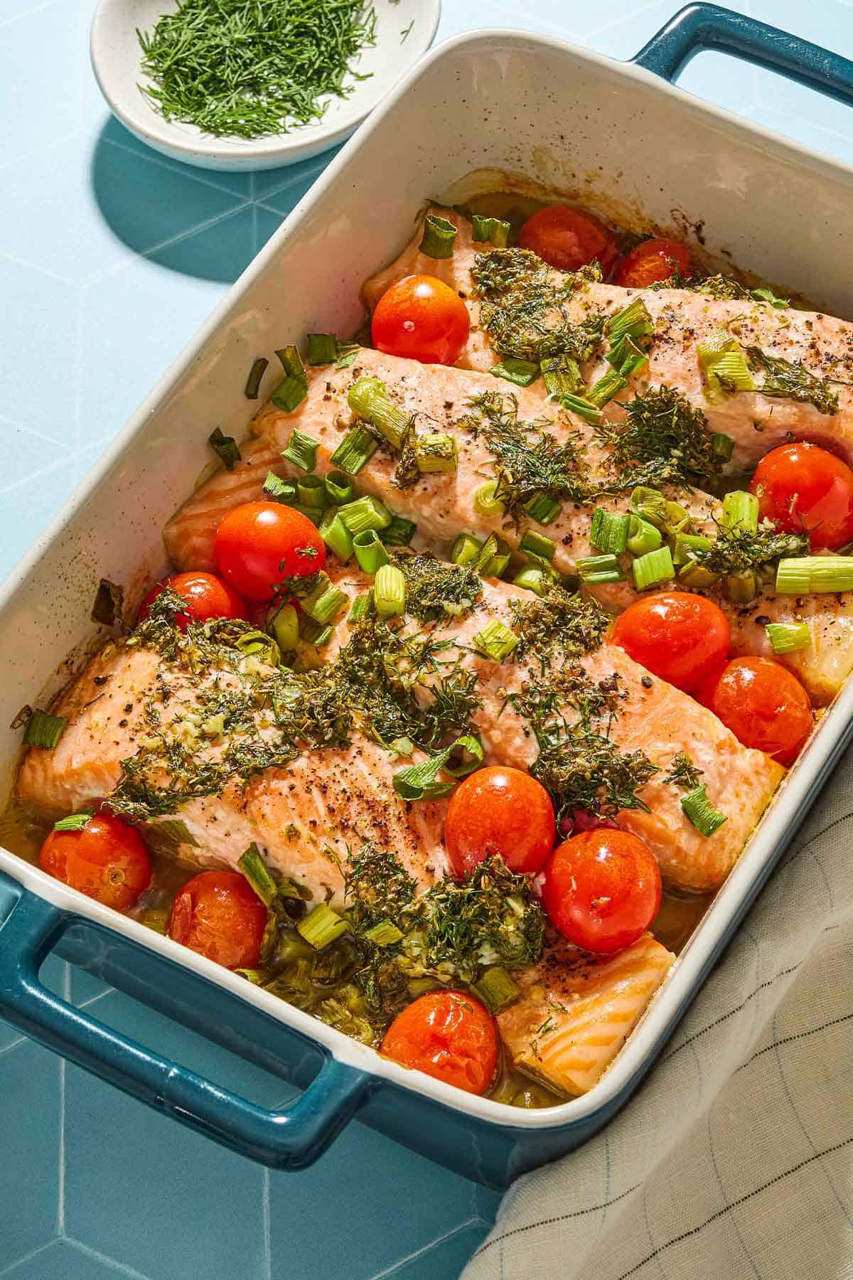 4 filets de saumon grec aux tomates cerises dans un plat allant au four. À côté se trouve un petit bol d’aneth.