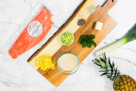 Fond en marbre blanc avec une planche à découper en bois recouverte des ingrédients de cette recette avec un filet de saumon, un ananas et un poireau entourant la planche à découper