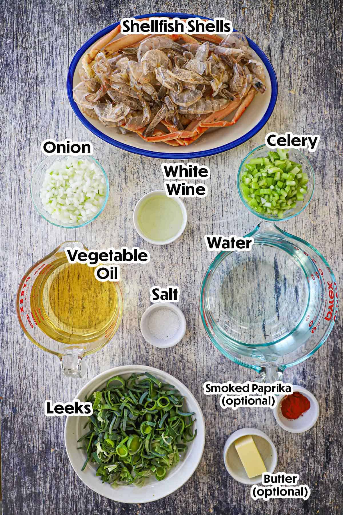 Un arrangement d'ingrédients pour un bouillon de coquillages fait maison sur fond de bois gris comprenant des coquilles de coquillages, de l'oignon, du céleri, du vin, des poireaux, de l'huile et des assaisonnements.