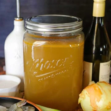 Une vue rapprochée d'un très grand pot en verre Mason rempli de bouillon de crustacés fait maison et entouré d'un bol rempli de crevettes crues, de poireaux, d'oignons et d'une bouteille de vin blanc.