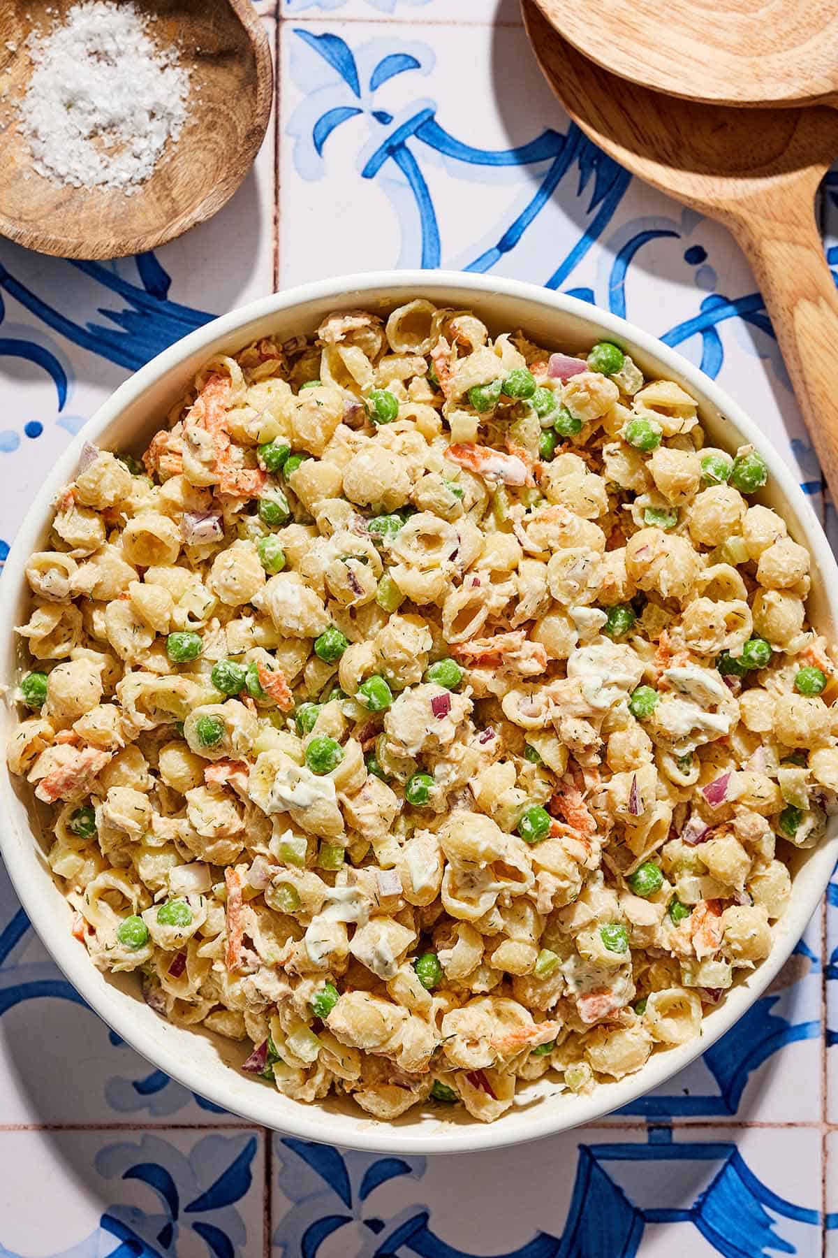 Salade de pâtes au thon dans un bol de service à côté d'un bol de sel et d'ustensiles de service en bois.