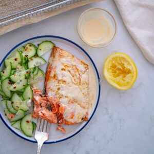 air fryer frozen salmon recipe in plate next to air fryer basket with cucumber salad