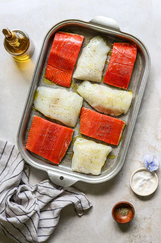Le saumon rouge et le poisson blanc assaisonnés de sel, de poivre et d'huile d'olive trônent sur un plat d'accompagnement dans cette recette parfaite pour griller les plats de fête du 4 juillet.