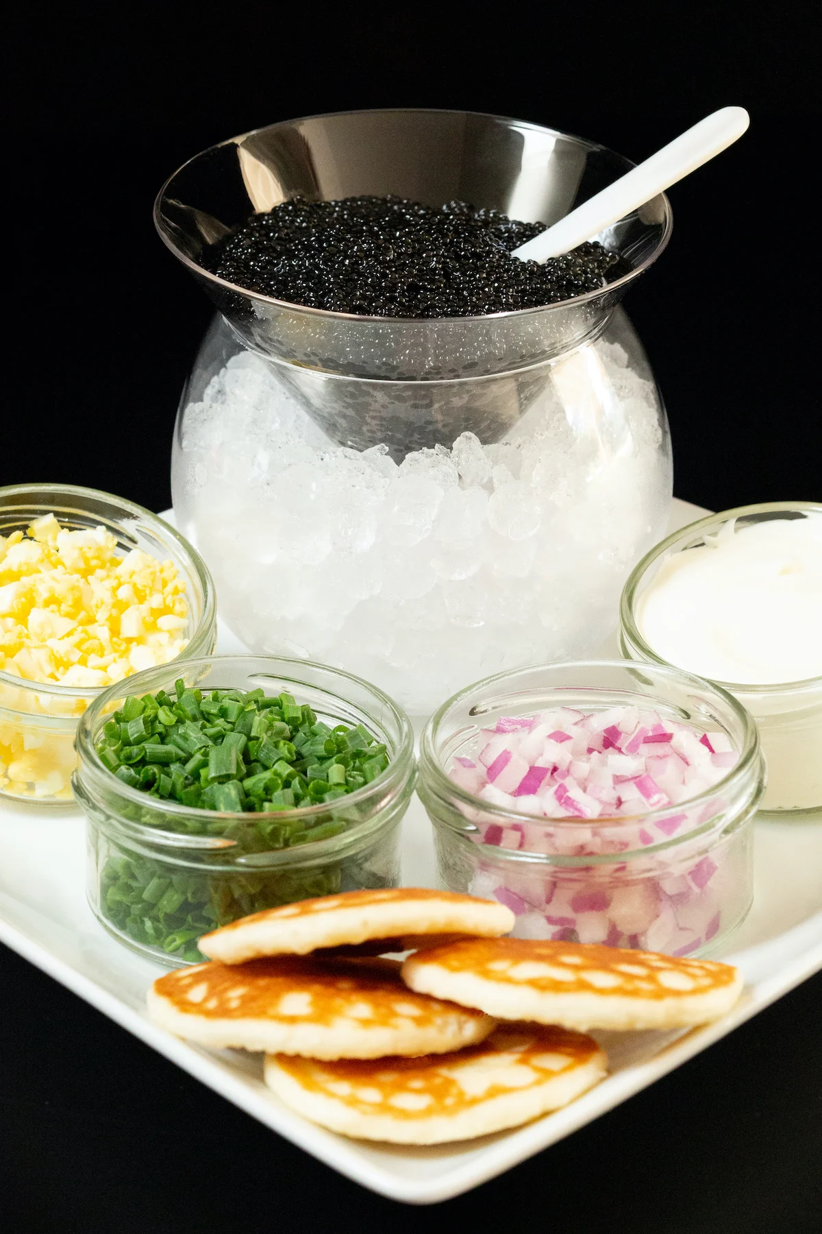 A glass cone is filled with caviar and sits on top of a glass bowl filled with ice. Around it are jars of chopped boiled egg, green onion, red onion, and creme fraiche as well as blinis.