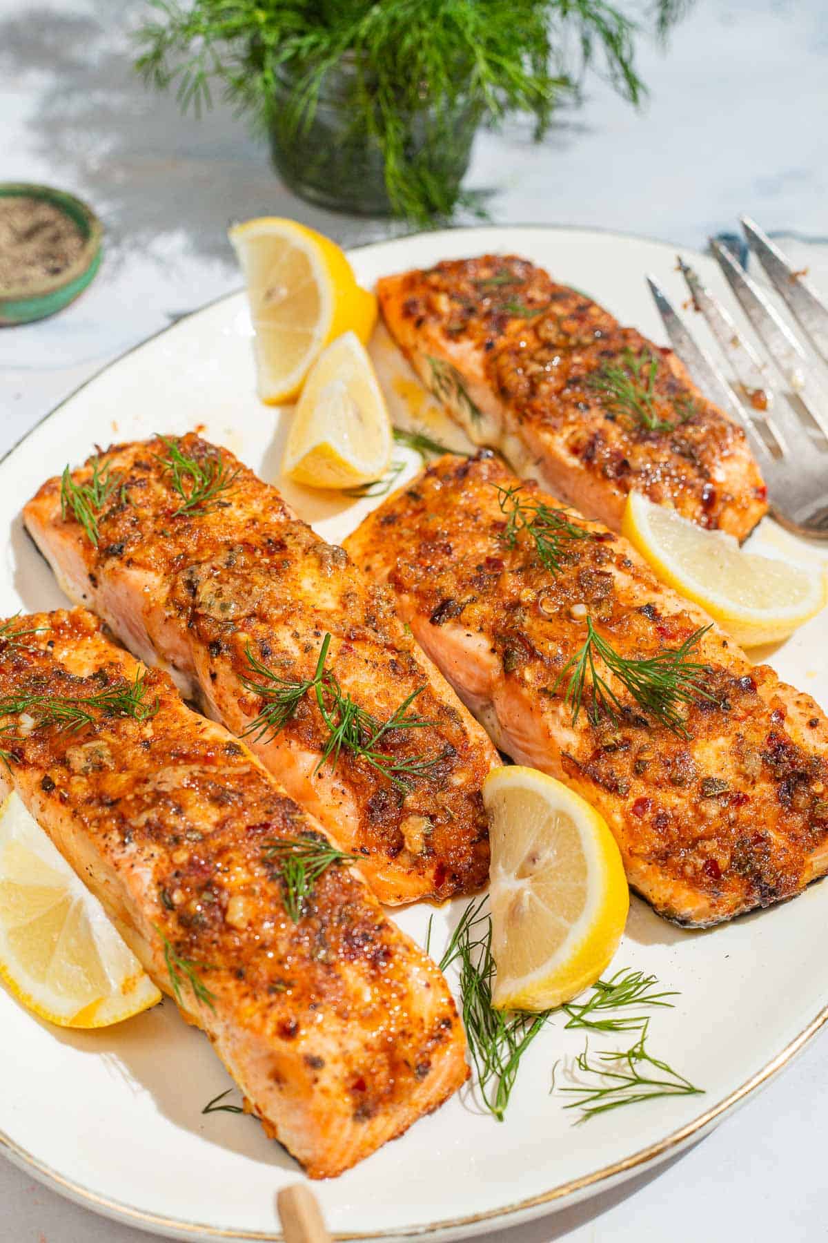 Un gros plan de 4 filets de saumon de Dijon cuits au four sur un plateau de service avec des quartiers de citron et une fourchette. À côté se trouvent un bol de poivre noir et un bouquet d’aneth.