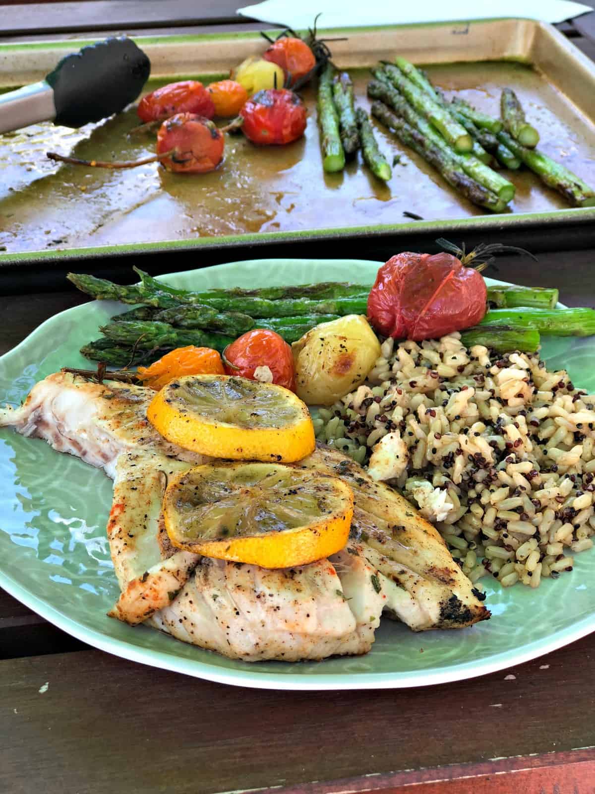 assiette de repas devant une plaque de cuisson avec du poisson grillé garni de rondelles de citron, de tomates raisins et d'asperges.