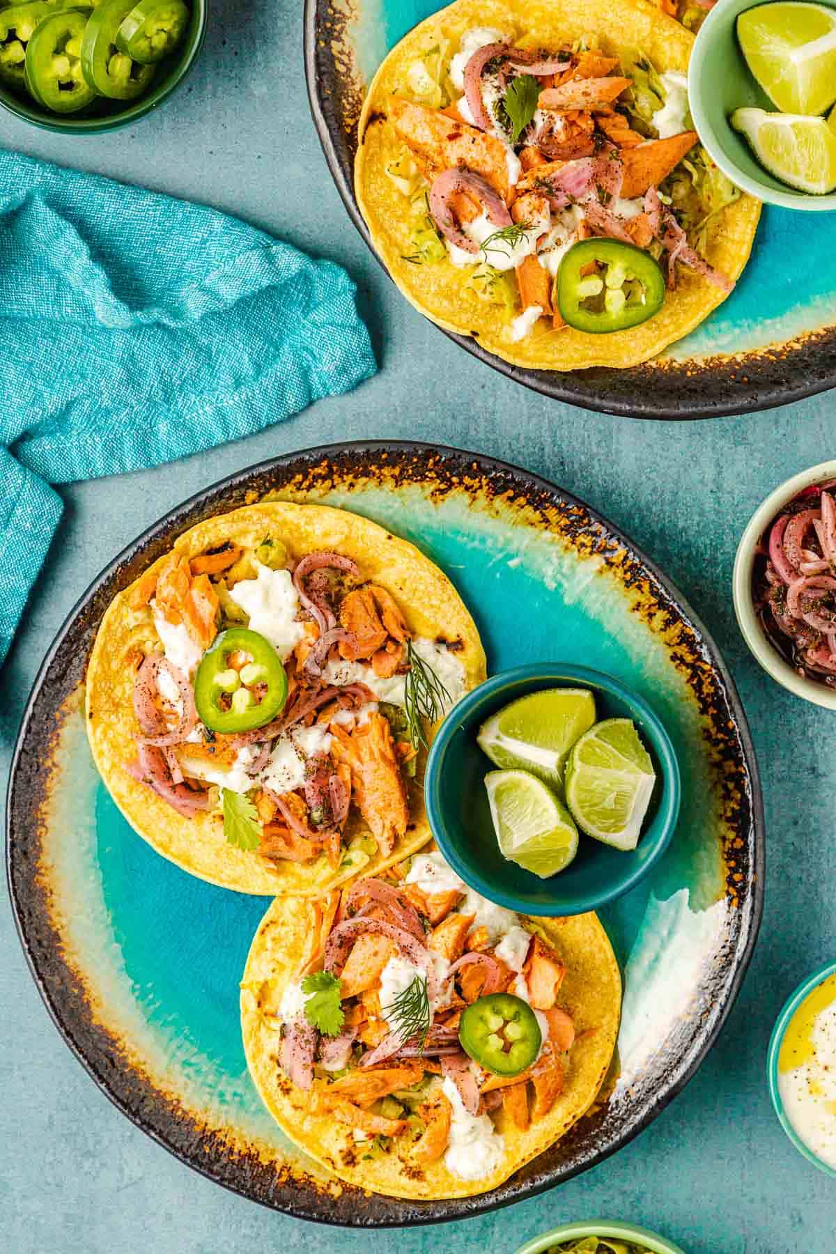 deux tacos au saumon ouverts sur des tortillas de maïs et un bol de 3 quartiers de citron vert sur une assiette, avec une autre assiette de tacos au saumon et un bol de jalapenos tranchés en arrière-plan.