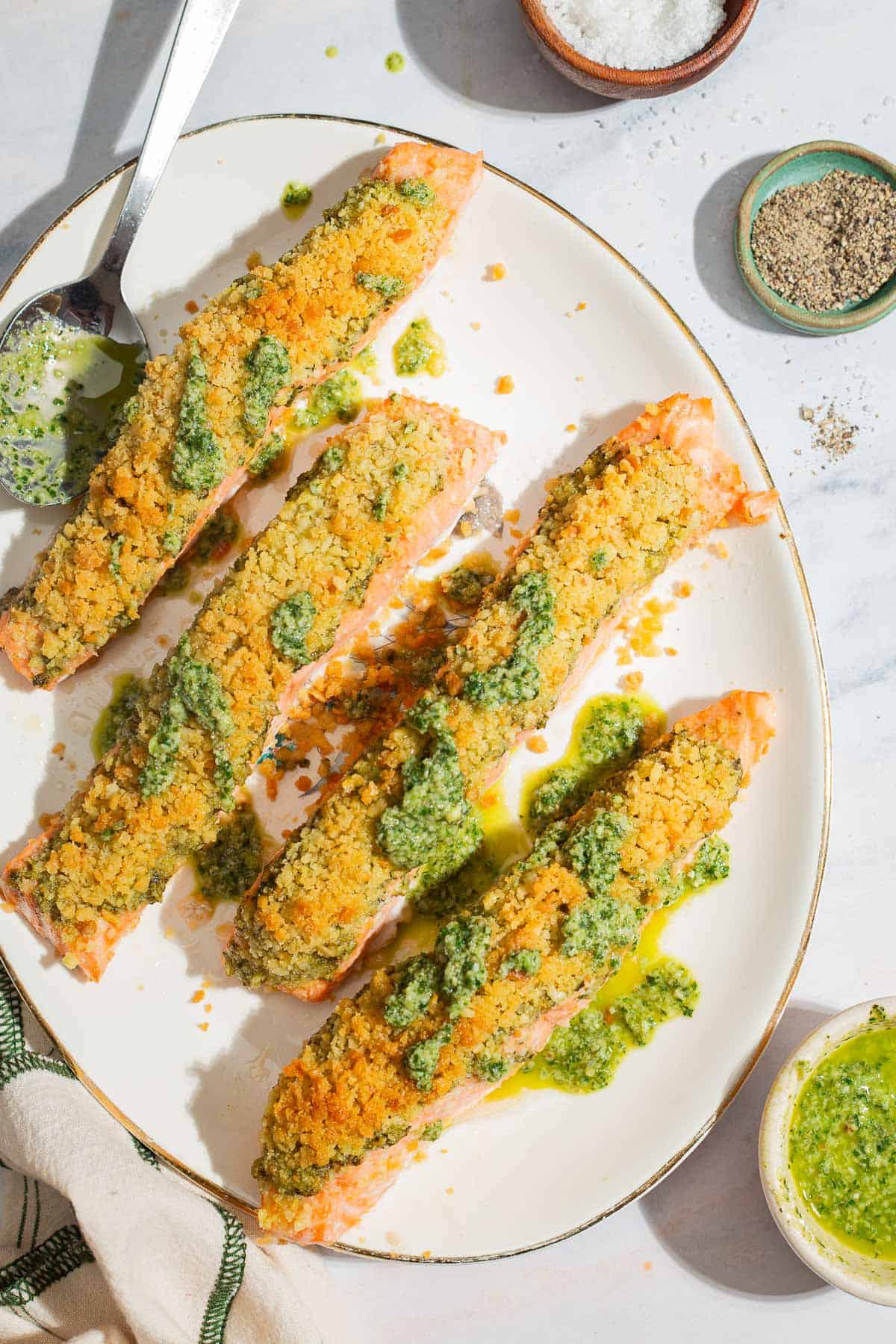 Une photo aérienne de 4 filets de saumon au pesto sur un plateau avec une cuillère. À côté se trouvent des bols de sel, de poivre et de pesto.