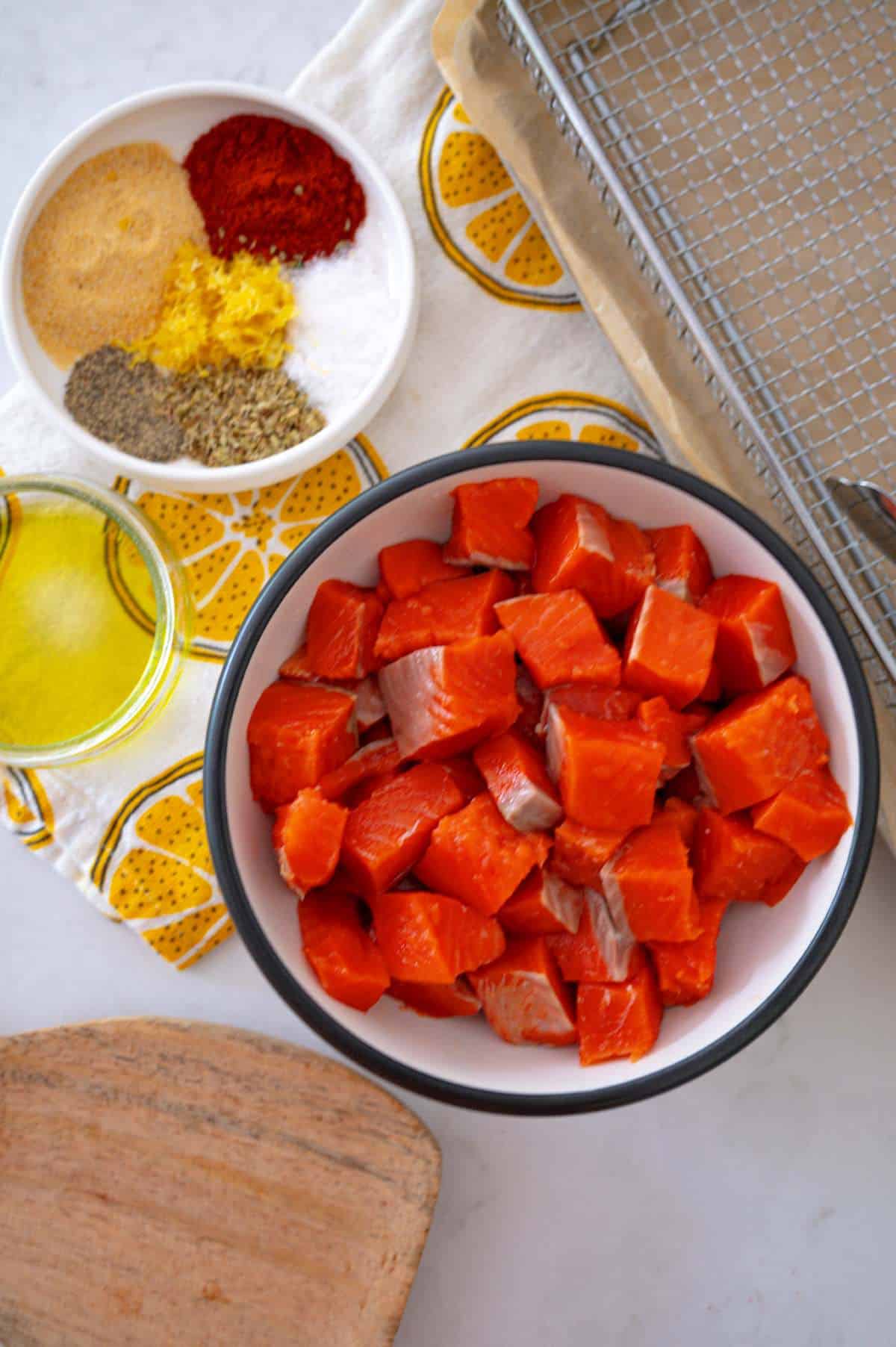 ingrédients pour faire des bouchées de saumon à la friteuse à air. bol de morceaux de saumon près d'une assiette d'épices et d'huile d'olive. serviette citronnée à proximité